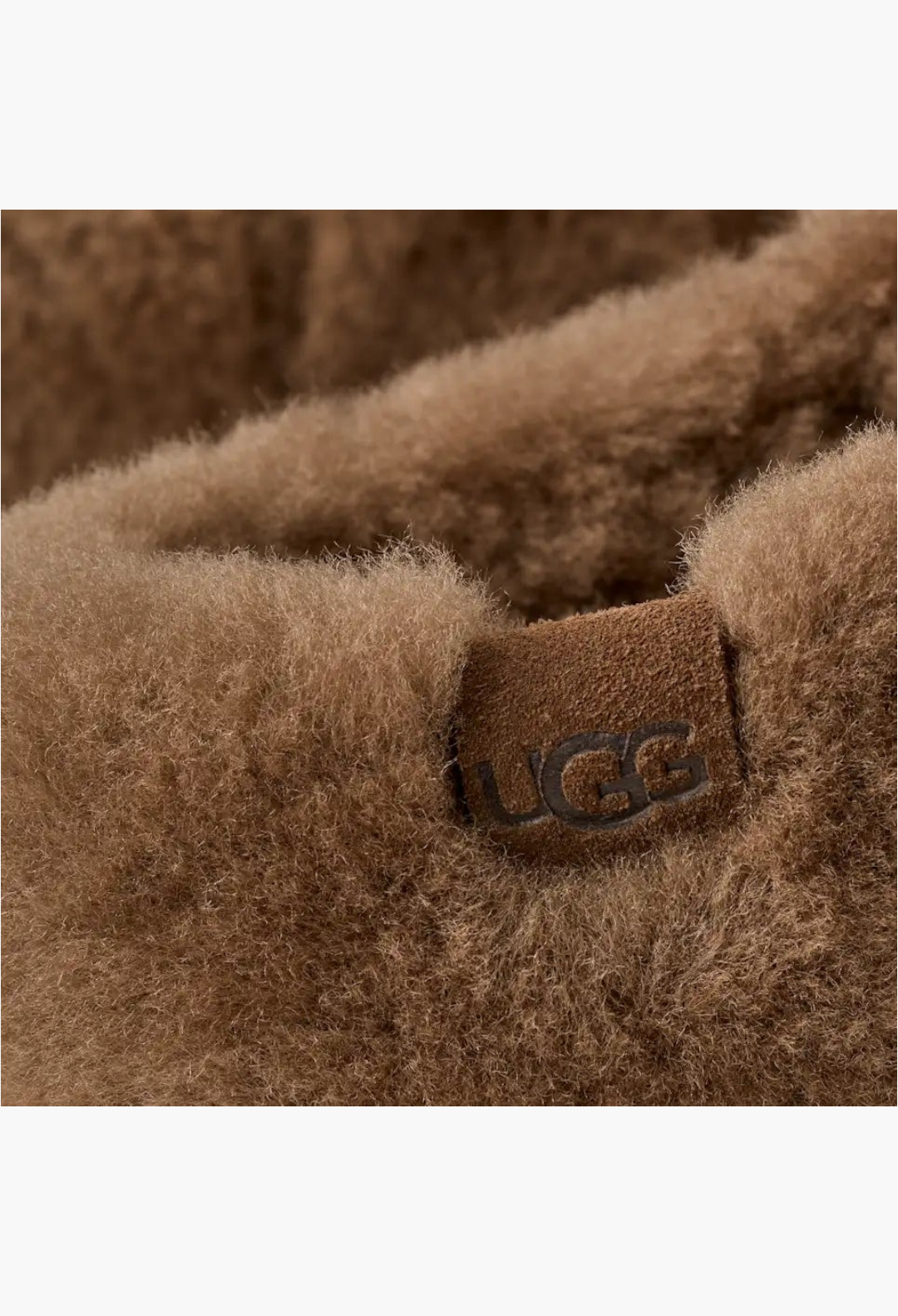 Close-up of GLAM MODA Elea Fluffed Slip-On featuring soft brown sheepskin texture and an embossed UGG logo suede tag.