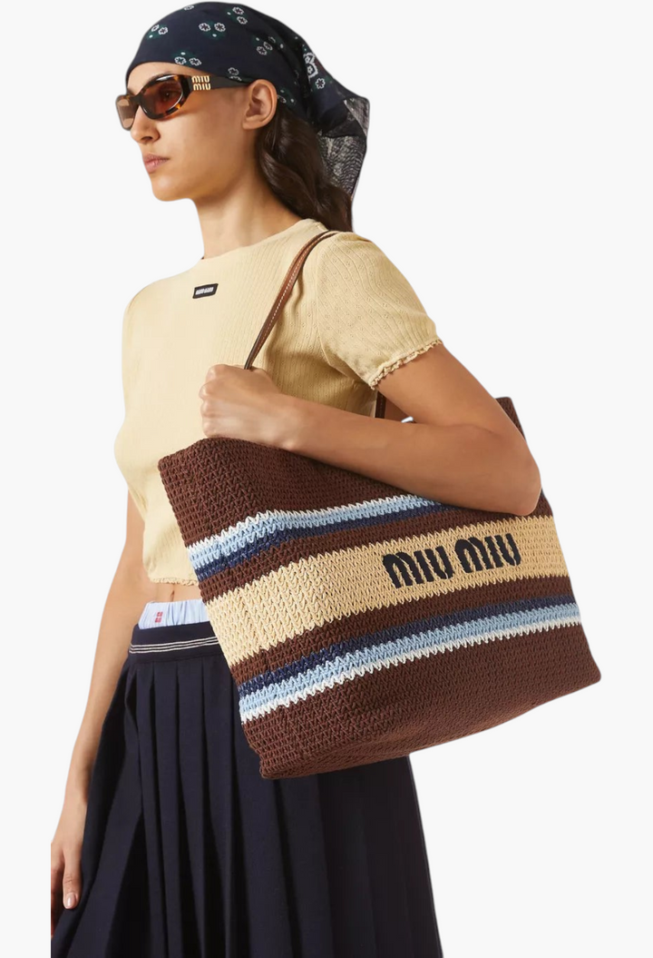 GLAM MODA brown raffia-effect tote bag with Miu Miu logo, leather handles, and striped pattern worn by a model.