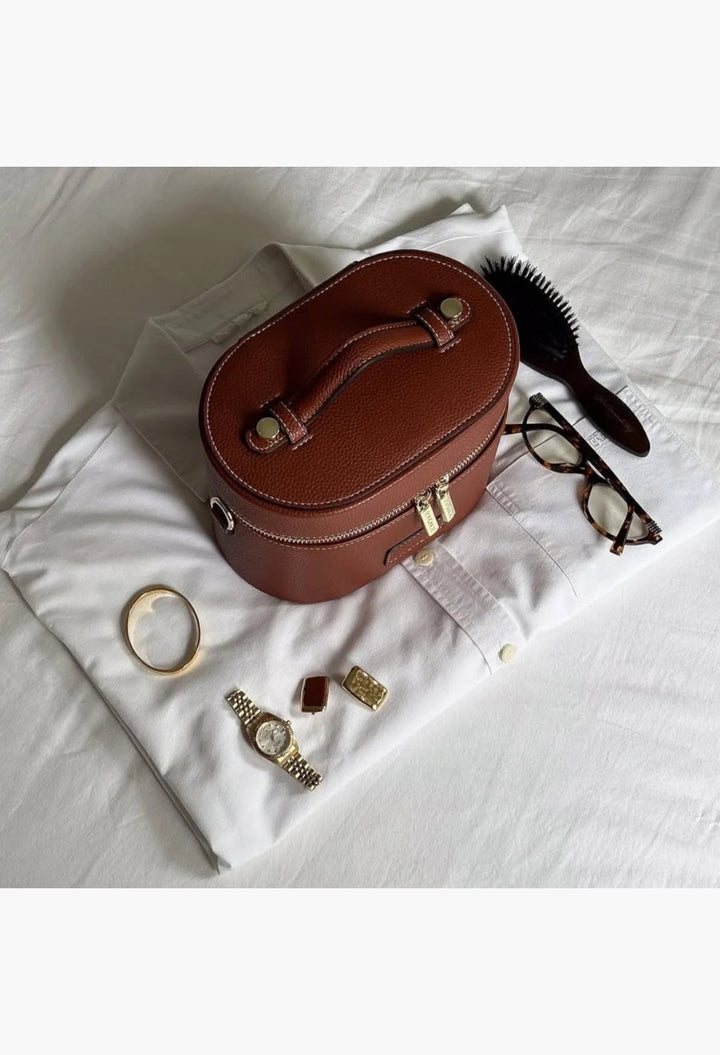GLAM MODA brown vegan leather oval toiletry case with a top handle and gold zippers, displayed on a white shirt.