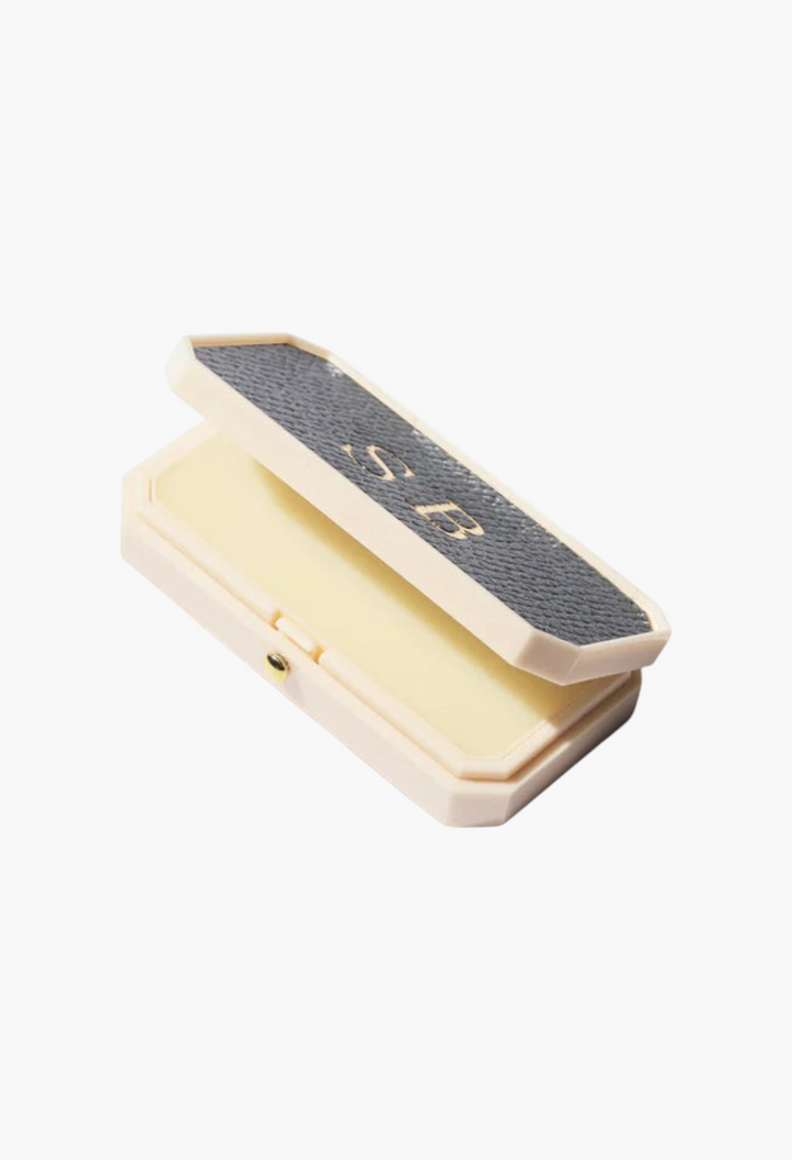 GLAM MODA Baume des Muses Ivory lip balm in a vintage-style cream case with a black leather-like lid and gold initials.