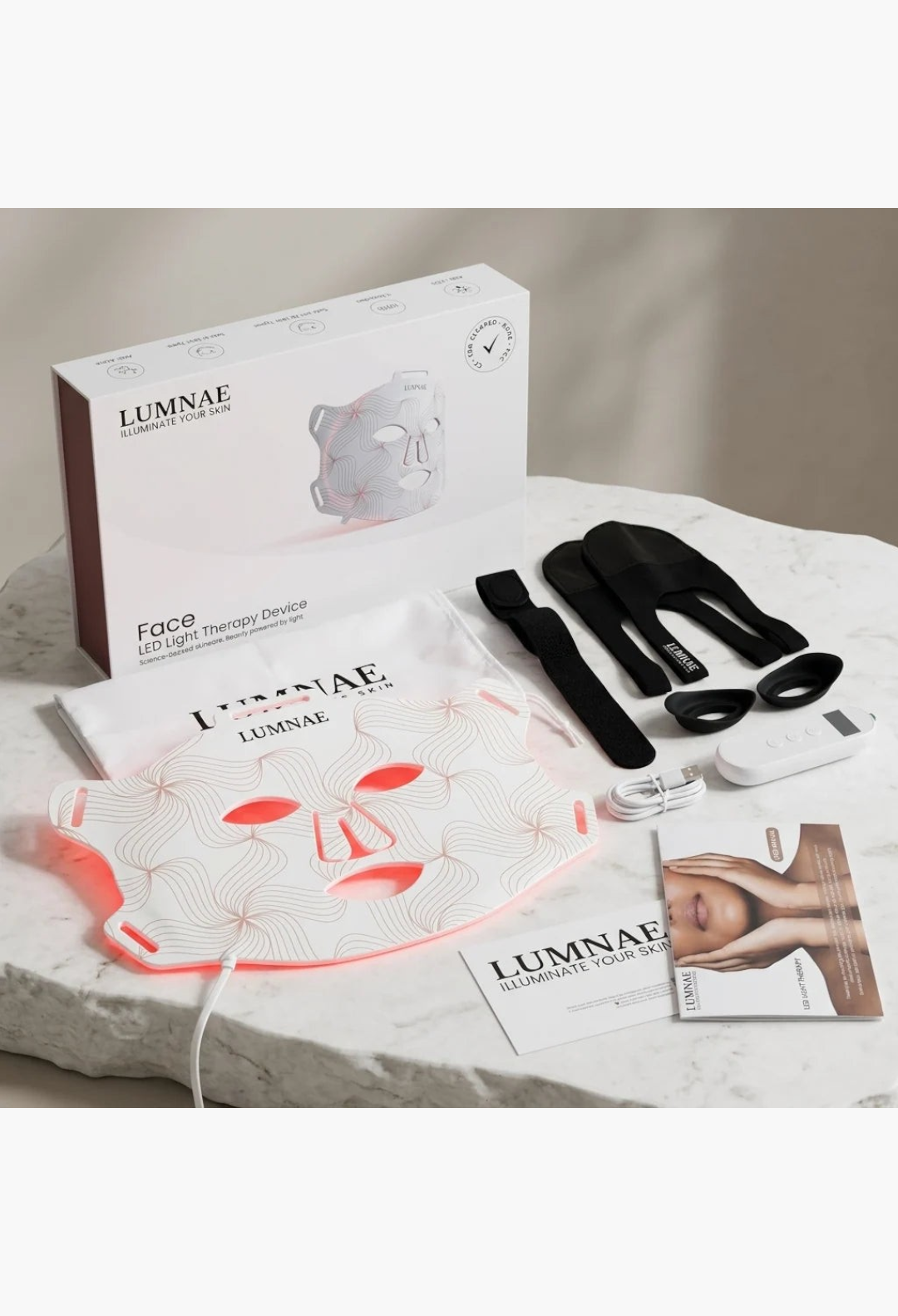 GLAM MODA Lumnae LED light therapy mask kit with silicone mask, remote, straps, and packaging on a marble surface.