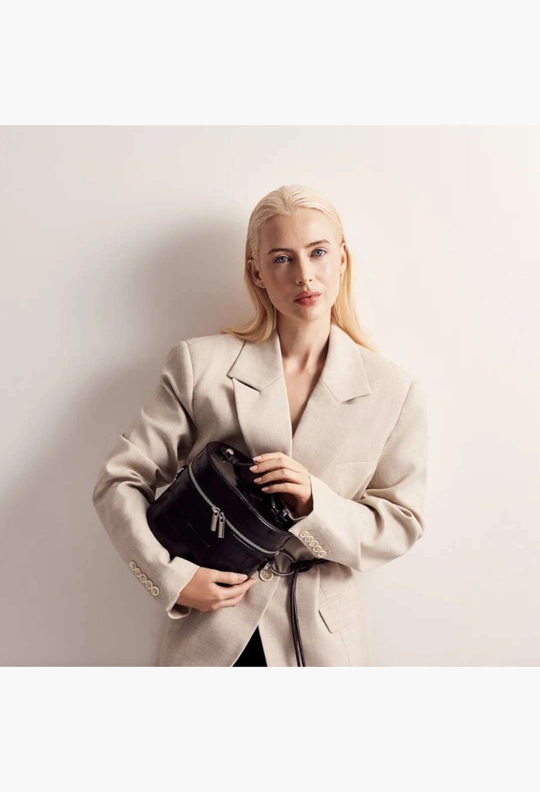 GLAM MODA oval black vegan leather toiletry case held by a blonde model wearing a beige blazer
