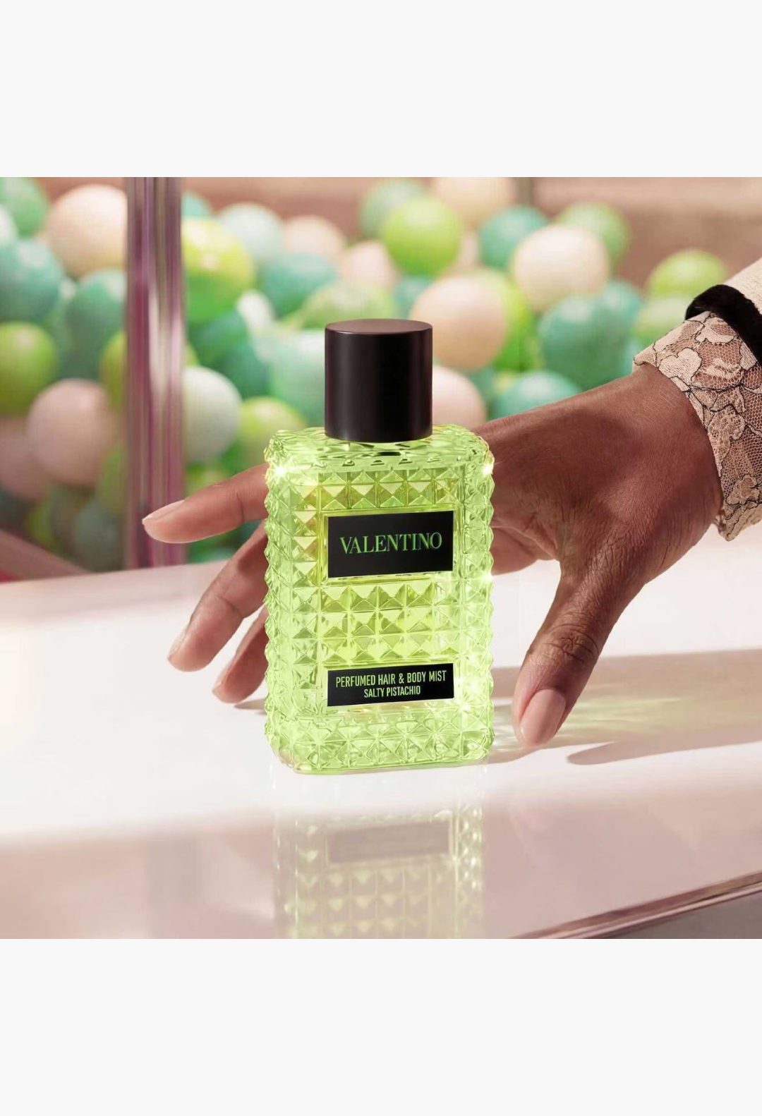 GLAM MODA Valentino Born in Roma Salty Pistachio hair and body mist in a green studded glass bottle held by a hand.