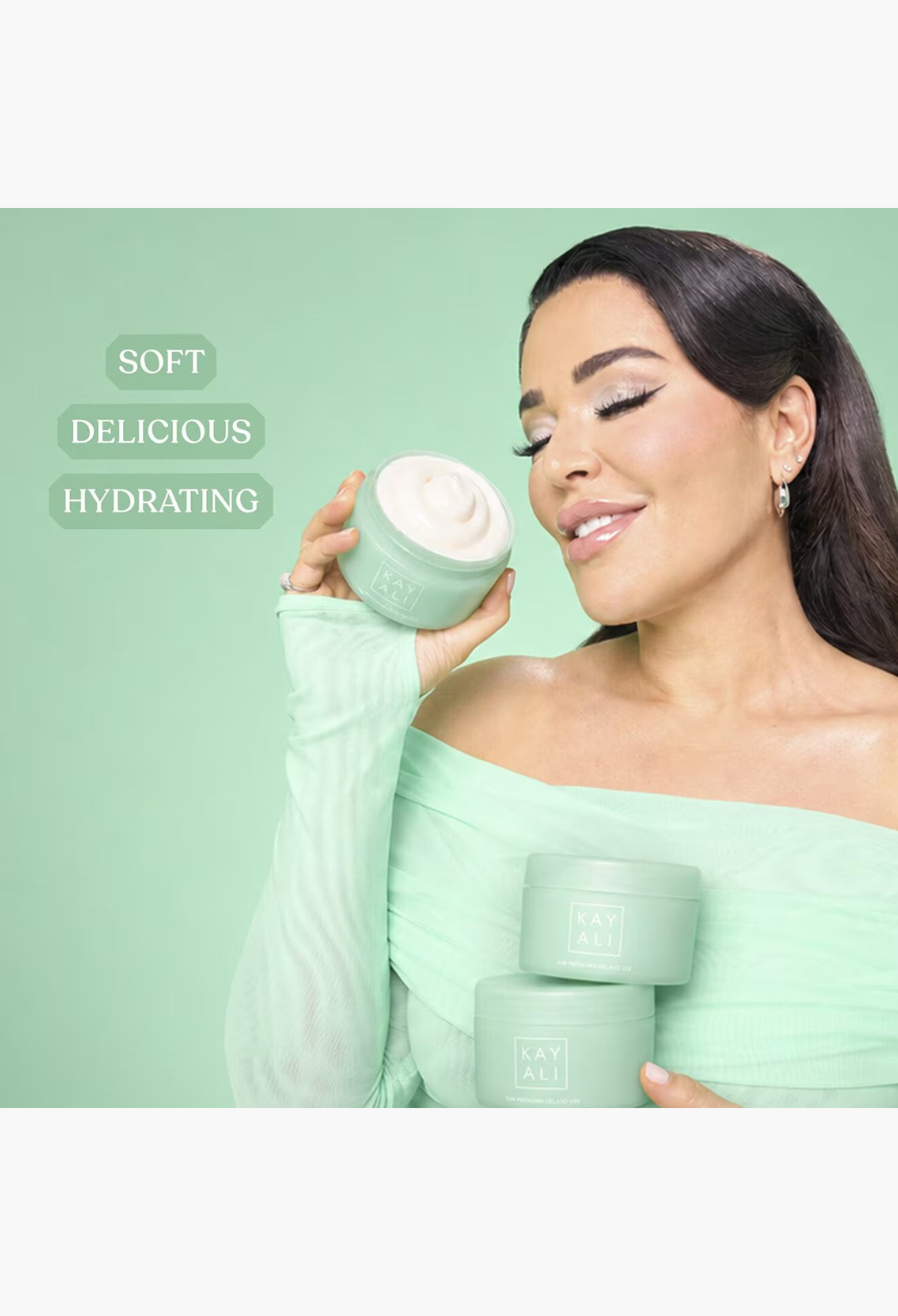 GLAM MODA model holding KAYALI Yum Pistachio Silk Soufflé body cream jars against a soft green background.