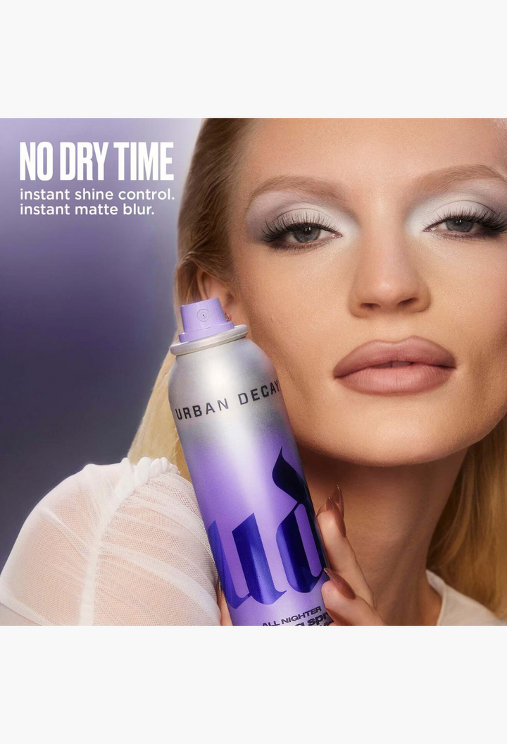 GLAM MODA Ghost Spray setting powder in a purple gradient aerosol bottle held by a model with matte makeup.