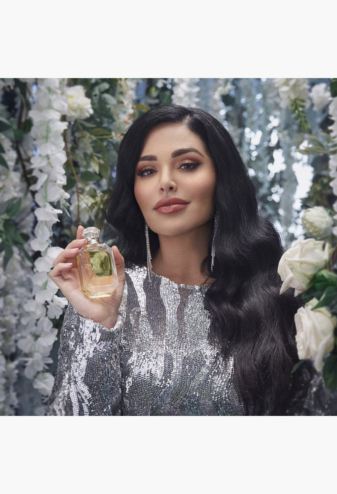 GLAM MODA model holding a glass bottle of Kayali Déjà Vu White Flower 57 perfume with a silver sequined dress