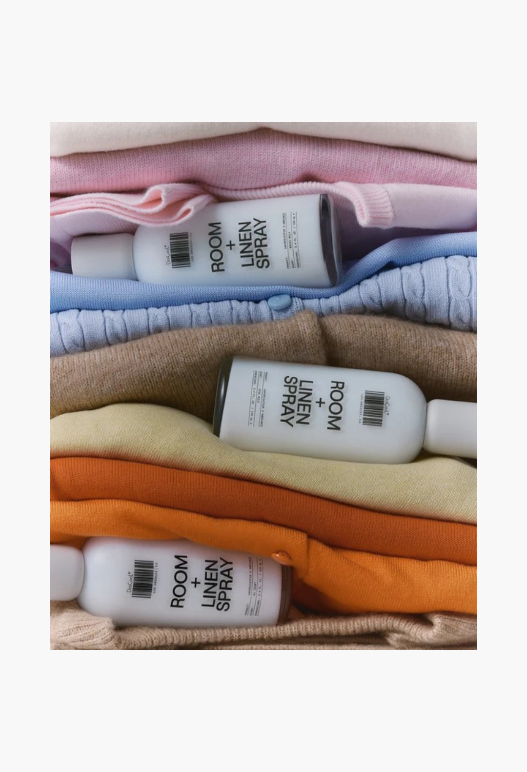 GLAM MODA Room + Linen Spray bottles in 01 Taunt resting on a stack of folded pastel and warm-toned knit sweaters.