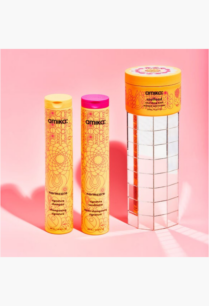 GLAM MODA Amika hair care set featuring Normcore shampoo, conditioner, and Soulfood Nourishing Mask on a mirrored stand.