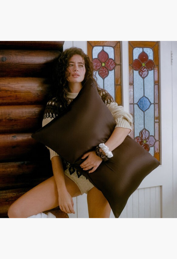 GLAM MODA chocolate brown pure silk queen pillowcase held by a woman in a cozy sweater against a rustic log wall
