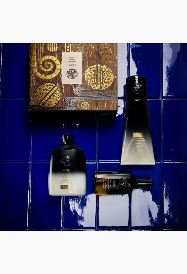GLAM MODA Oribe Gold Lust haircare set featuring shampoo, conditioner, and nourishing oil on a blue tiled background.