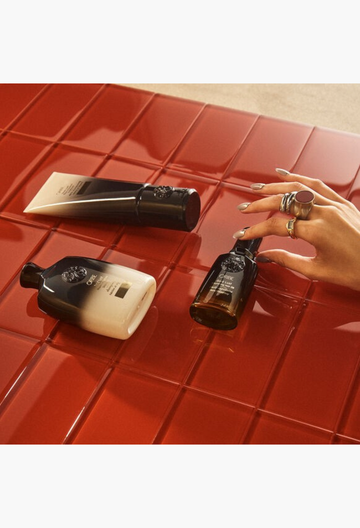 GLAM MODA Oribe Gold Lust haircare set featuring shampoo, conditioner, and nourishing oil on a glossy red tiled surface.