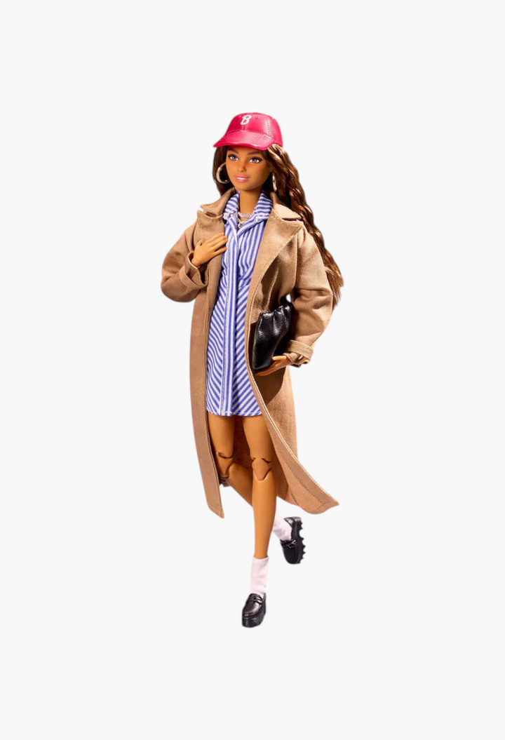 GLAM MODA New York Fashion Week Barbie doll wearing a tan trench coat, striped shirt dress, and red baseball cap.
