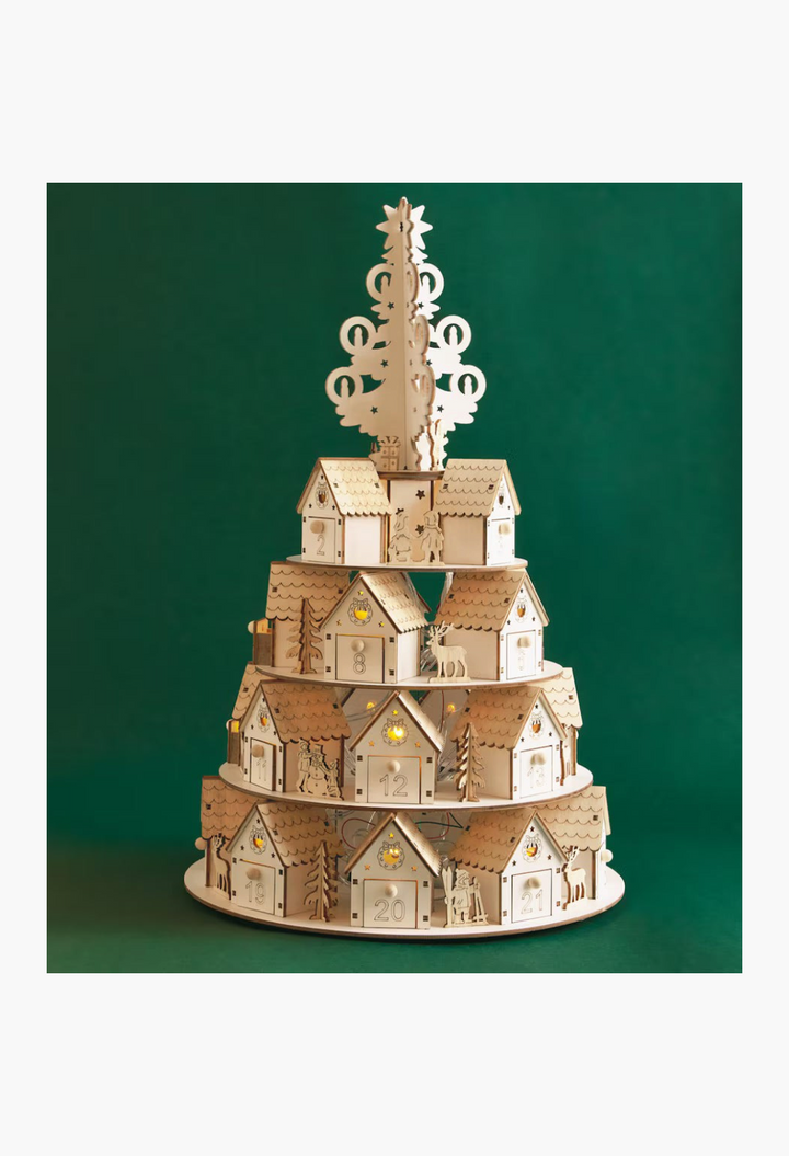 GLAM MODA tiered wooden 3D advent calendar featuring miniature houses and a Christmas tree topper on a green background