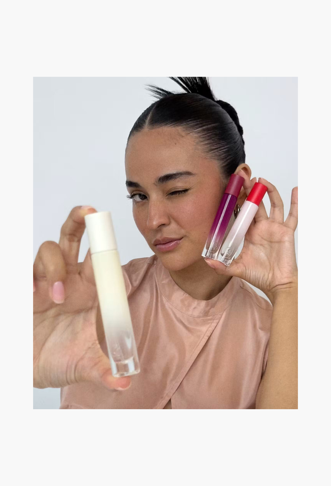 GLAM MODA model winking while holding Glossier You Doux woody perfume bottles in a studio setting