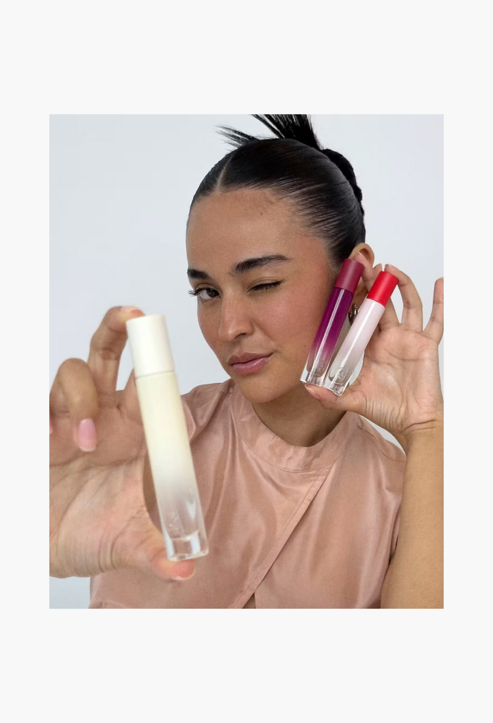 GLAM MODA model winking while holding Glossier You Doux woody perfume bottles in a studio setting