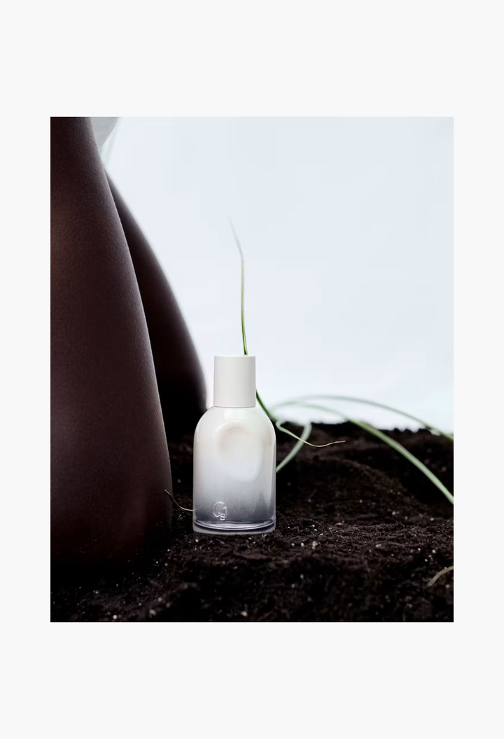 GLAM MODA Glossier You Doux eau de parfum in a white gradient bottle set in dark soil with a green blade of grass.