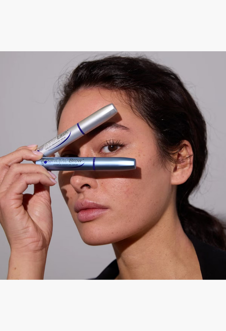 GLAM MODA model holding RapidLash and RapidBrow enhancing serum tubes near her eyes against a white background