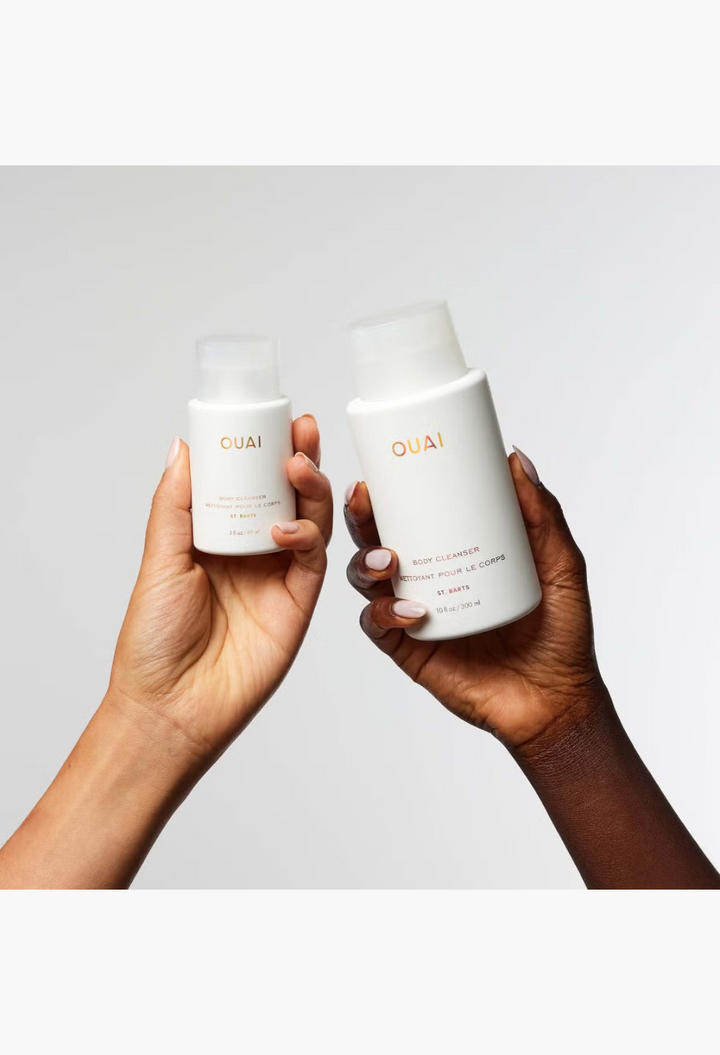 GLAM MODA OUAI St. Barts Body Cleanser bottles held in two hands against a plain white background.