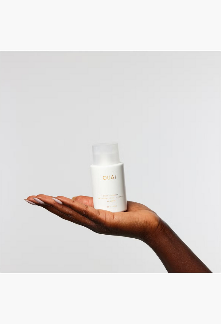 GLAM MODA hand holding a white bottle of OUAI St. Barts Body Cleanser against a plain light background