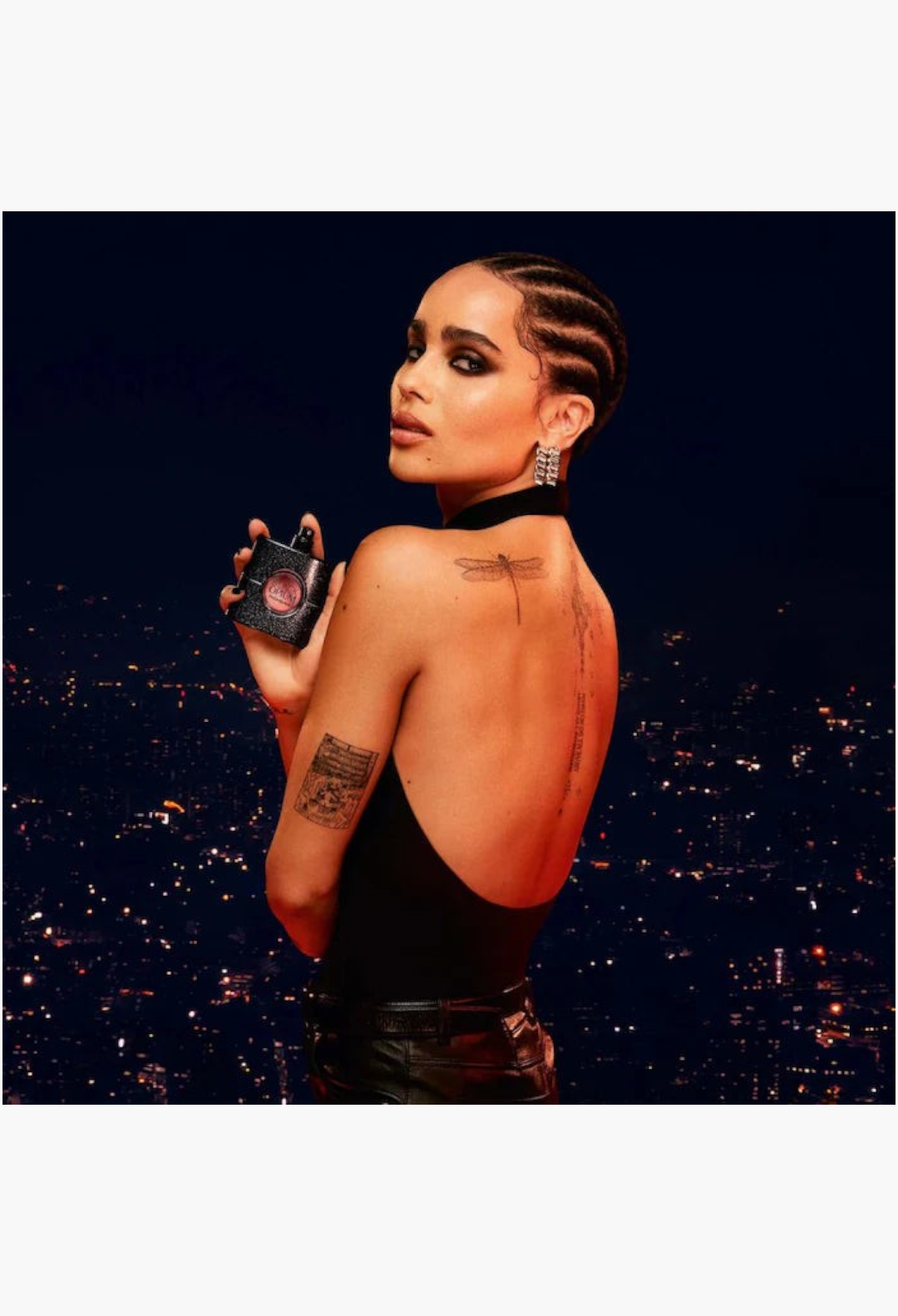 GLAM MODA Black Opium perfume bottle held by a woman with dark hair and tattoos against a city night background