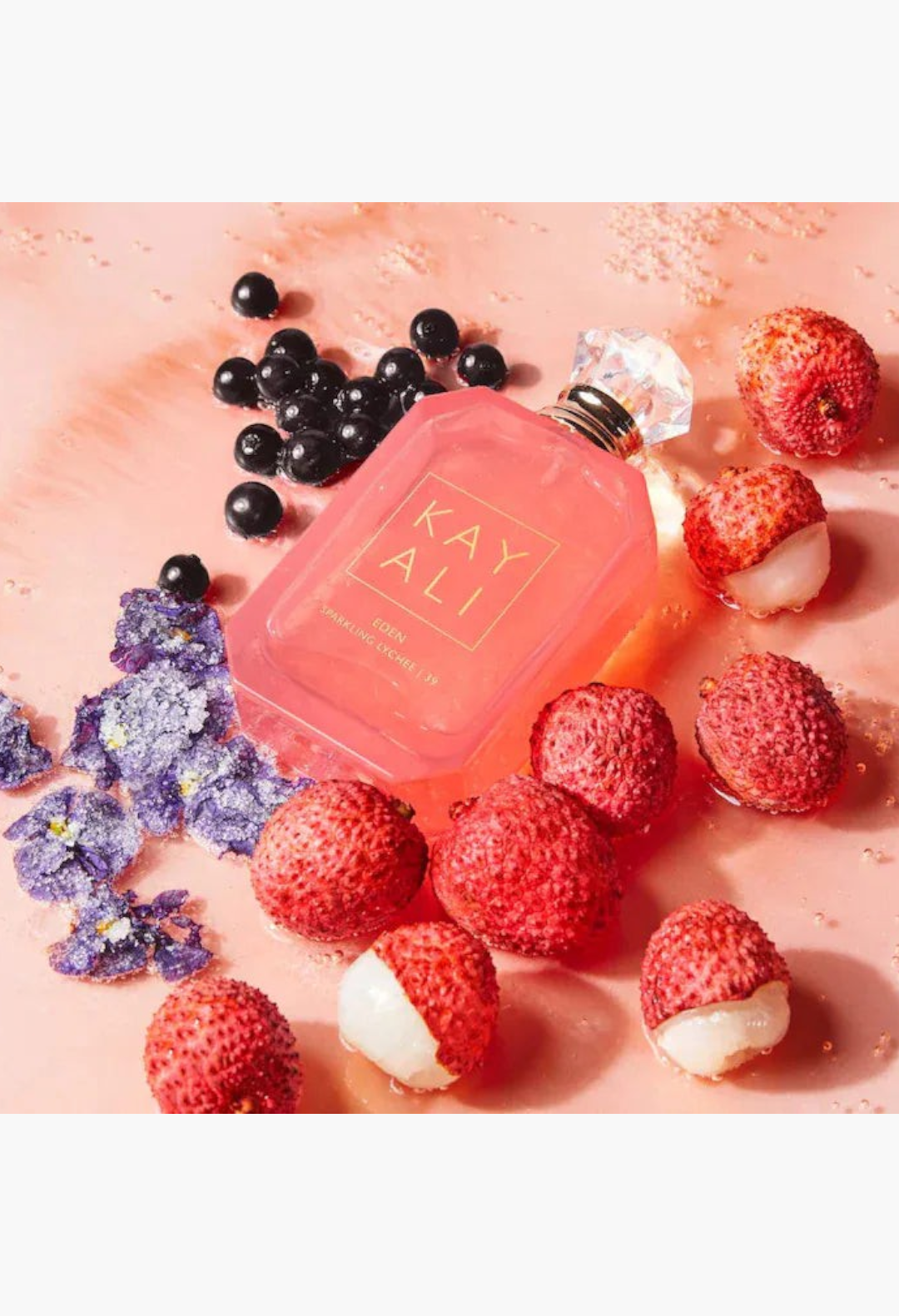 GLAM MODA Kayali Eden Sparkling Lychee perfume bottle surrounded by fresh lychee fruit and purple floral accents.