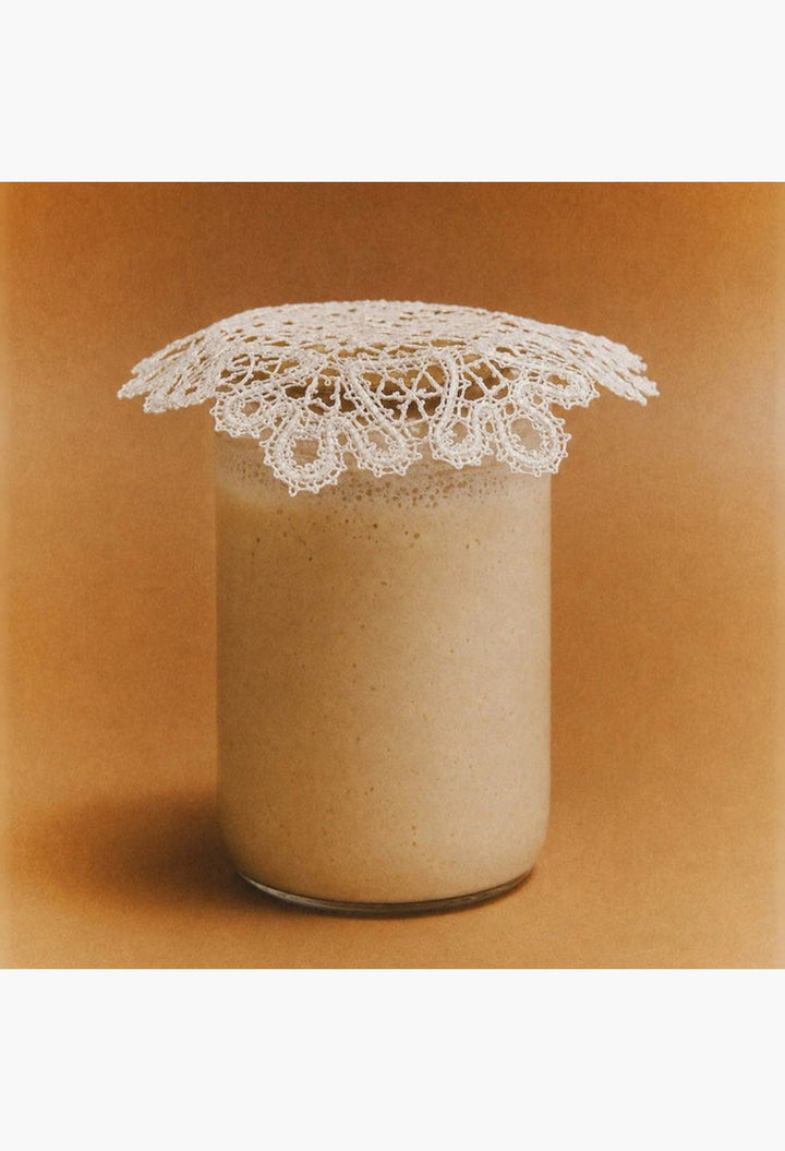 GLAM MODA Le Chouchou peptide lip balm in a clear jar topped with a delicate white lace doily on a tan background.