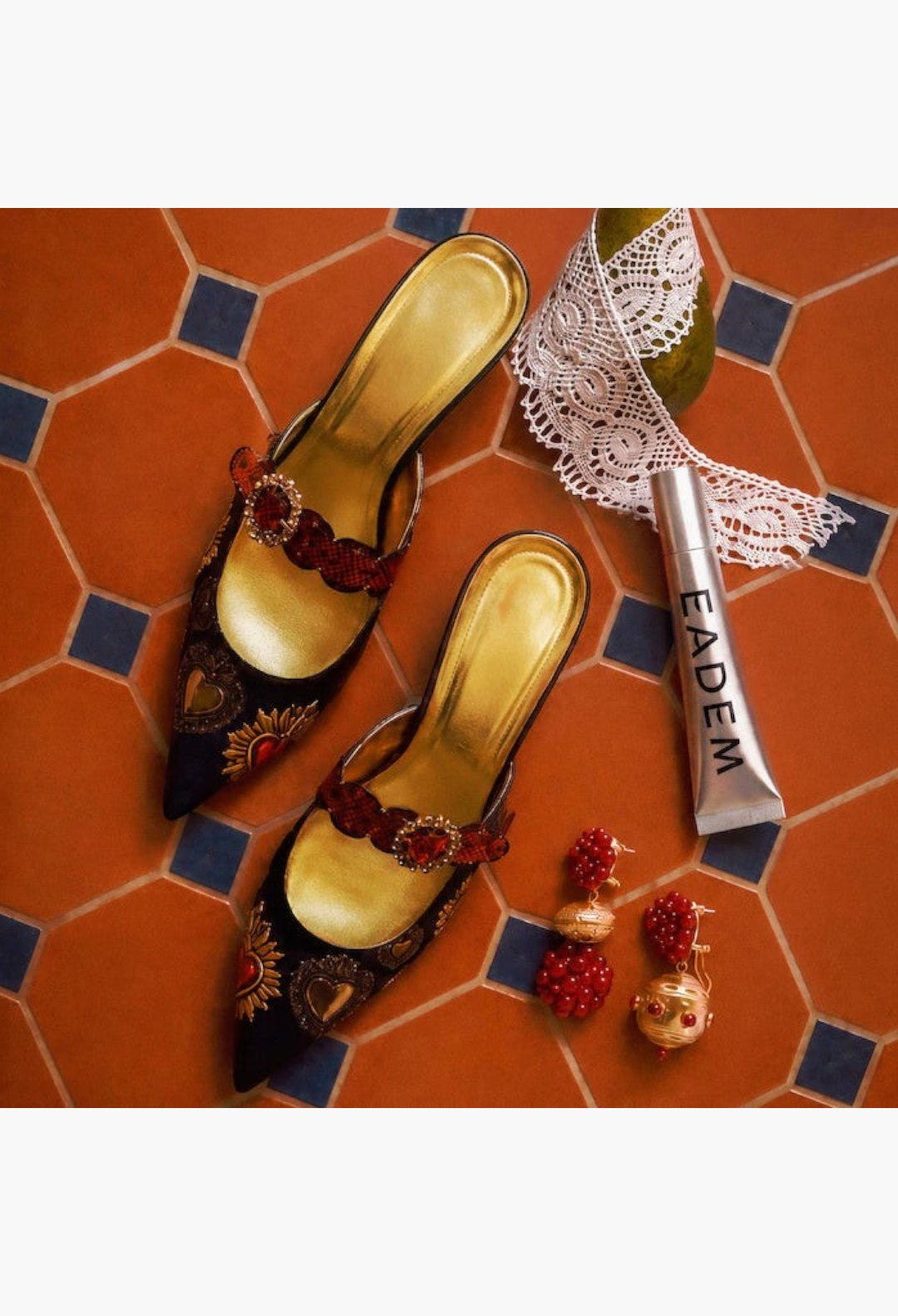 GLAM MODA Le Chouchou peptide lip balm tube next to ornate black pointed heels, lace, and red beaded earrings.