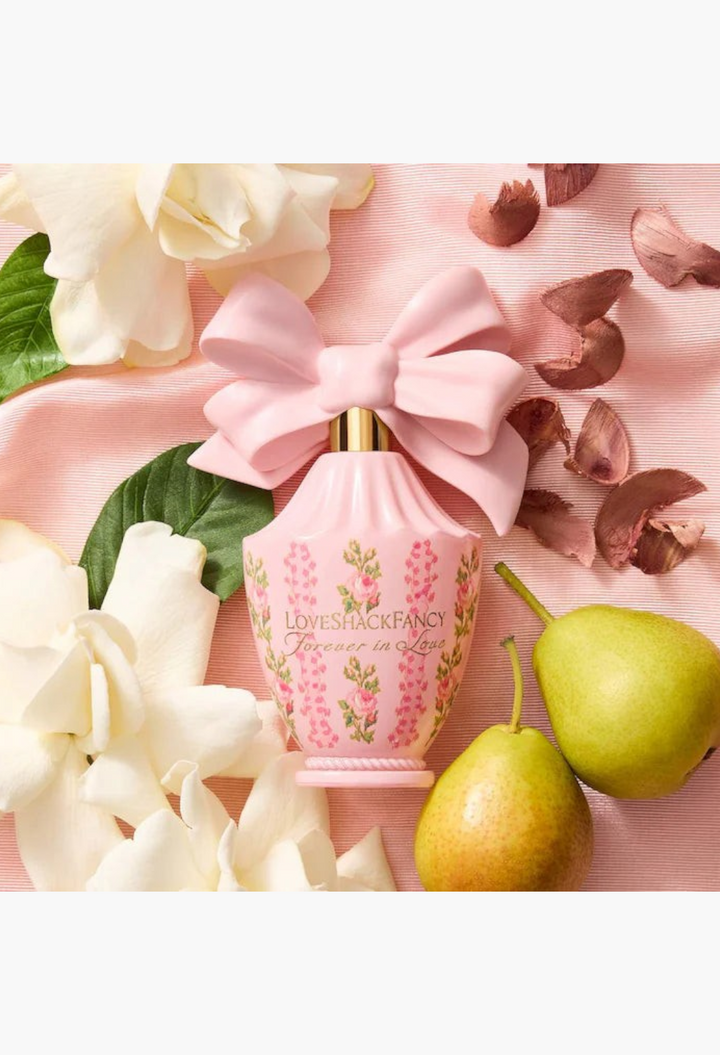 GLAM MODA LoveShackFancy Forever In Love perfume bottle with a pink bow cap, surrounded by white flowers and pears.